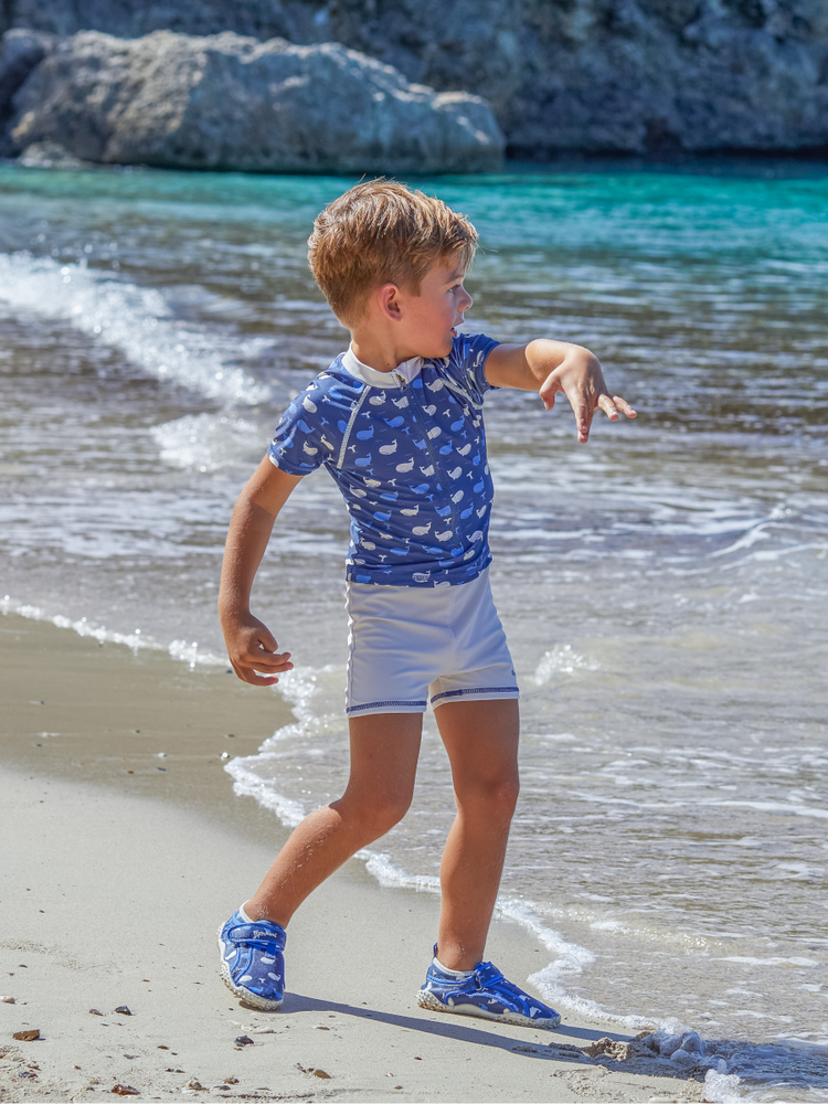 Junge am Strand in blauer UV-Badebekleidung und Playshoes Aquaschuhen mit Wellenmuster – idealer Sonnenschutz und rutschfeste Badeschuhe für aktive Kinder. Perfekte Strandmode und Kinderschuhe für Wasser und Sand – erhältlich bei meine-kinderschuhe.de.