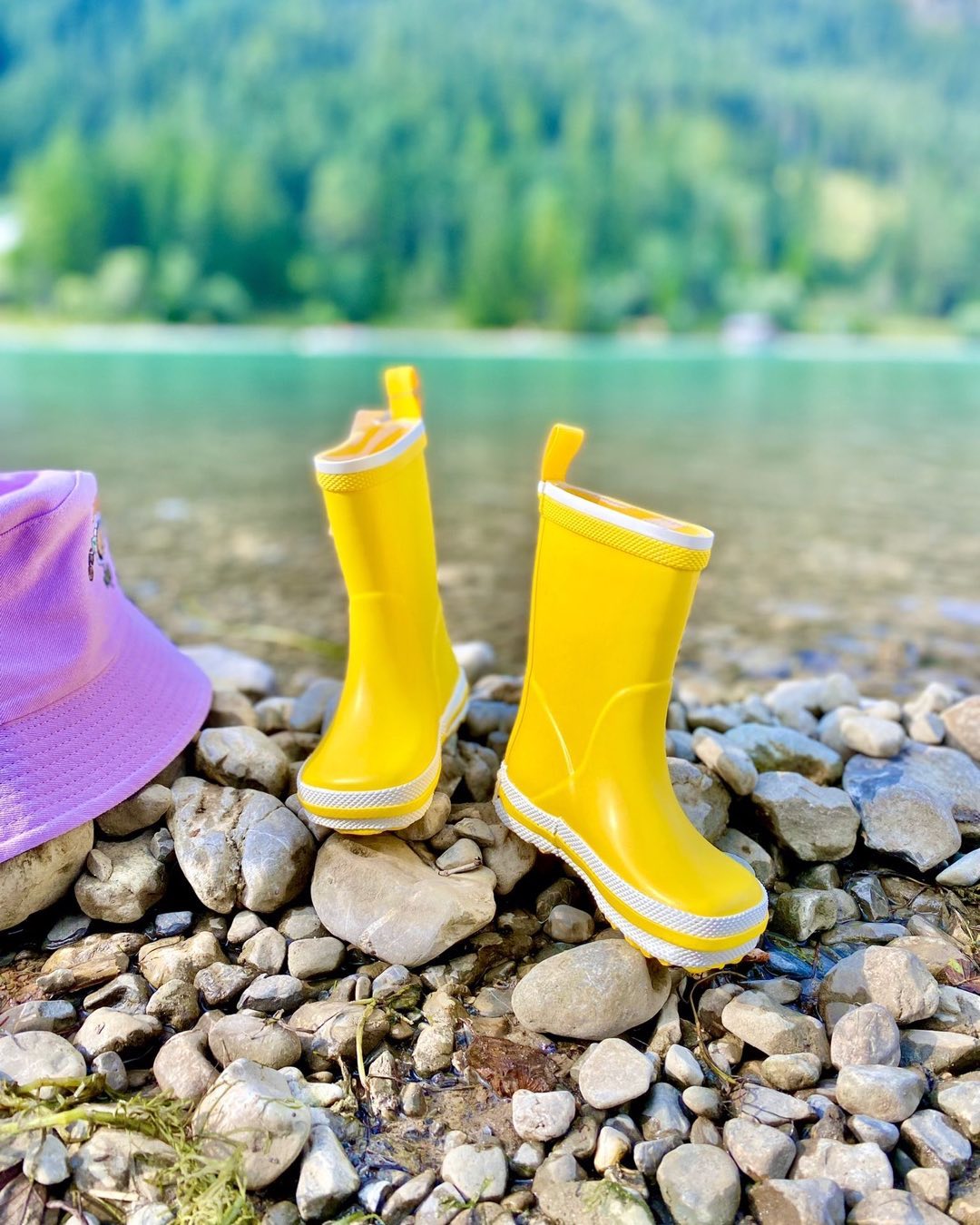 Gelbe Gummistiefel von der Marke Bundgaard auf Steinen mit einem Lila Hut und als Hintergrund ein grüner See